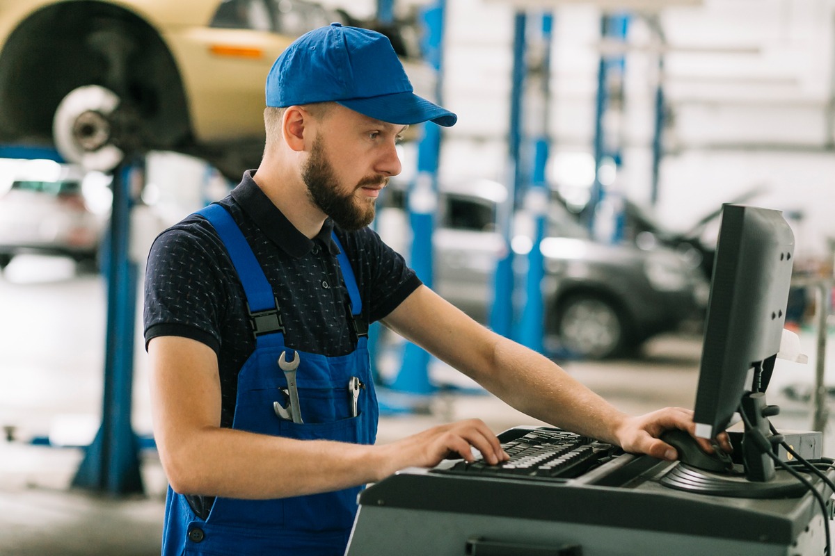 Mecânico de boné e macacão azul consultando o computador da oficina, com carros suspensos em elevadores ao fundo