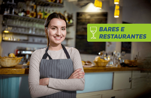 Atendente de avental sorrindo de braços cruzados no salão de um restaurante, com balcão e cestas de pão ao fundo e o selo Bares e Restaurantes